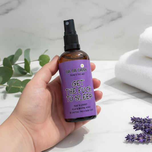 Hand holding a bottle labeled 'Get the F*** to Sleep' with lavender flowers and towels in the background.