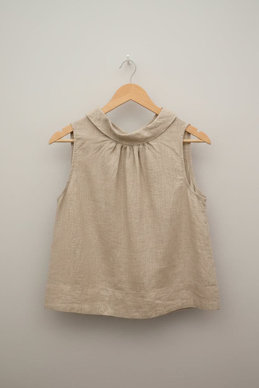 A sleeveless, high-neck linen top in a light beige color, hanging on a wooden hanger against a white paneled wall.